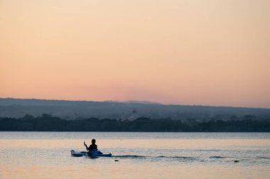 Huzurlu suda sessizce kürek çeken bir kanocu, uzak tepelerle sakin günbatımının tadını çıkarır..