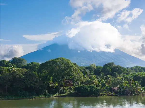 Yüksek volkan kısmen bulutlarla örtülmüş, canlı yeşillik ve mavi gökyüzünün altında sakin bir nehir tarafından kucaklanmış..