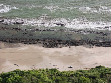 Kumlu sahil açık gökyüzünün altında uzanırken yumuşak dalgalar kayalara çarpıyor..