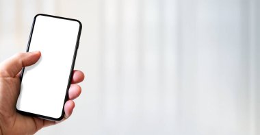 Hand holding a smartphone with blank white screen. White office background. Horizontal banner.
