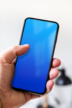 Hand holding a smartphone with blank blue screen. White office background.