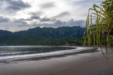 Marquesas Adaları 'nın Nuku Hiva kentindeki Taiohae Körfezi' nin siyah kumsal sahili yemyeşil bitki örtüsü, hindistan cevizi ağaçları ve bulutlu bir gökyüzünün altındaki engebeli yeşil dağlar tarafından çerçevelenmiştir. Fransız Polinezyası