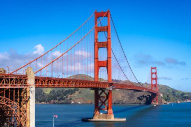 Golden Gate Köprüsü körfez boyunca uzanır, kırmızı kuleleri mavi gökyüzü ve su ile zıtlaşır, arka planda yeşil tepeler vardır.