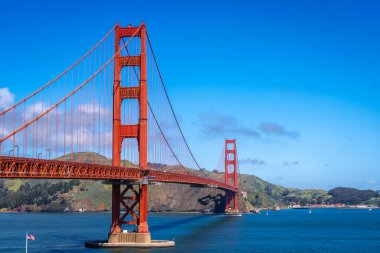 Golden Gate Köprüsü körfez boyunca uzanır, kırmızı kuleleri mavi gökyüzü ve su ile zıtlaşır, arka planda yeşil tepeler vardır.