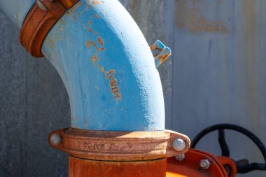 Paslanmış eklemleri ve metal vanaları olan eskimiş mavi ve turuncu endüstriyel su boruları karmaşık bir ağ oluşturur. Aşınmış metal yüzeyler kentsel arkaplan ile tezat oluşturuyor
