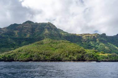 Marquesas Adaları, Fransız Polinezyası 'ndaki Tahuata Adası' nda engebeli yeşil dağlar, sarp kayalıklar, yemyeşil bitki örtüsü ve dağınık bulutlu gökyüzünün altında mavi okyanus suları bulunur.