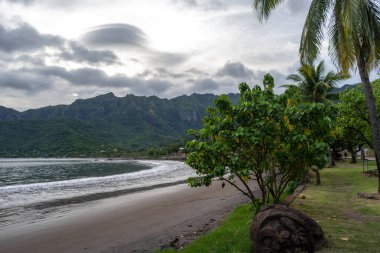 Marquesas Adaları 'nın Nuku Hiva kentindeki Taiohae Körfezi' nin siyah kumsal sahili yemyeşil bitki örtüsü, hindistan cevizi ağaçları ve bulutlu bir gökyüzünün altındaki engebeli yeşil dağlar tarafından çerçevelenmiştir. Fransız Polinezyası