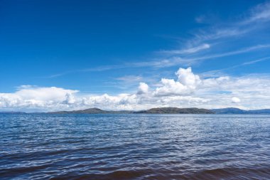 Llachon, Peru 'dan Titicaca Gölü' nün huzurlu manzarası. Derin mavi suları, dağınık adaları, parlak gökyüzünün altında yuvarlanan tepeleri ve tüylü bulutları var.