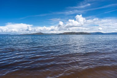 Llachon, Peru 'dan Titicaca Gölü' nün huzurlu manzarası. Derin mavi suları, dağınık adaları, parlak gökyüzünün altında yuvarlanan tepeleri ve tüylü bulutları var.