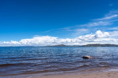 Peru, Titicaca Gölü kıyısındaki Llachon plajında sakin sular, altın kum ve tüylü bulutlarla dolu açık mavi bir gökyüzünün altında uzak bir dağ manzarası bulunur.