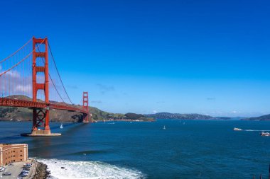 Golden Gate Köprüsü körfez boyunca uzanır, kırmızı kuleleri mavi gökyüzü ve su ile zıtlaşır, arka planda yeşil tepeler vardır.