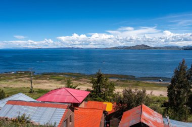 Llachon, Peru 'dan Titicaca Gölü' nün panoramik manzarası. Geleneksel kır evleri, yeşil alanlar ve dağınık bulutlu parlak bir gökyüzü altında sakin mavi sular.