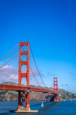 Golden Gate Köprüsü körfez boyunca uzanır, kırmızı kuleleri mavi gökyüzü ve su ile zıtlaşır, arka planda yeşil tepeler vardır.