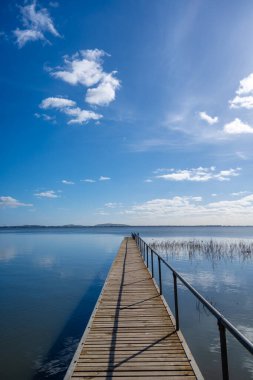 Victoria, Avustralya 'daki Colac Gölü' nün sakin suları üzerinde bir ahşap iskele uzanır. Mavi gökyüzü ve uzak tepeler huzurlu bir atmosfer yaratır.