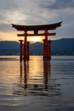 Itsukushima jinja Tapınağı 'nın ünlü yüzen torii kapısı Miyajima Adası yakınlarında sakin suları ve çevresini çevreleyen yeşil tepeleri olan günbatımında. Japonya, Hiroşima yakınlarında.