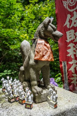 Kamakura 'daki Sasuke İnari-jinja ormanlarında minyatür adaklar taşıyan taş bir İnari tilki heykeli, canlı sancakların yanında duruyor.