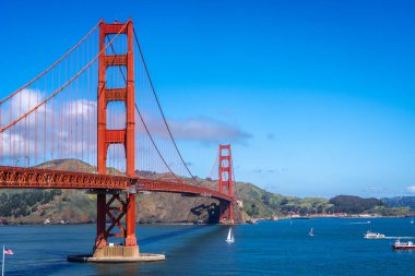 Golden Gate Köprüsü körfez boyunca uzanır, kırmızı kuleleri mavi gökyüzü ve su ile zıtlaşır, arka planda yeşil tepeler vardır.