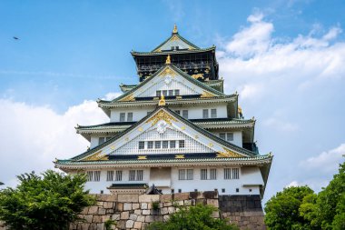 Osaka Kalesi 'nin görkemli ana kulesi parlak bir gökyüzü altında yeşil ağaçların üzerinde yükseliyor. Geleneksel Japon mimarisi ve altın süslemeleri sergiliyor.