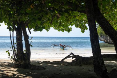Geleneksel ahşap bir tekne, Lia sahilinin beyaz kumları yakınında, Waleakodi Adası, Togian takımadaları, Sulawesi, Endonezya 'da parlak mavi bir gökyüzünün altında yüzmektedir.