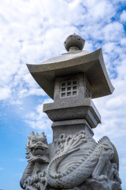 Japonya 'nın Enoshima adasındaki parlak bir gökyüzünün altında, efsanevi sanatla geleneksel tasarımı harmanlayan bir ejderha heykeli bulunan süslemeli taş fener.