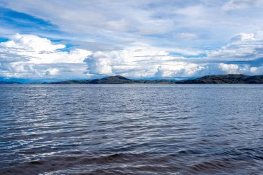 Llachon, Peru 'dan Titicaca Gölü' nün huzurlu manzarası. Derin mavi suları, dağınık adaları, parlak gökyüzünün altında yuvarlanan tepeleri ve tüylü bulutları var.