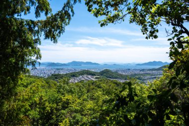 Hiroşima şehri ve Mitaki Dağı 'ndan Seto İç Denizi üzerindeki panoramik manzara, yeşil orman ve uzak mavi dağ sıraları tarafından çerçevelenmiştir.