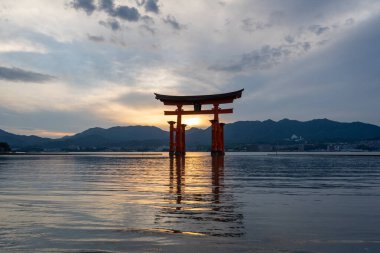 Itsukushima jinja Tapınağı 'nın ünlü yüzen torii kapısı Miyajima Adası yakınlarında sakin suları ve çevresini çevreleyen yeşil tepeleri olan günbatımında. Japonya, Hiroşima yakınlarında.