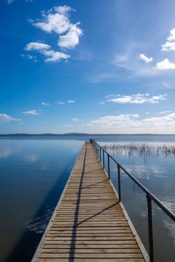 Victoria, Avustralya 'daki Colac Gölü' nün sakin suları üzerinde bir ahşap iskele uzanır. Mavi gökyüzü ve uzak tepeler huzurlu bir atmosfer yaratır.