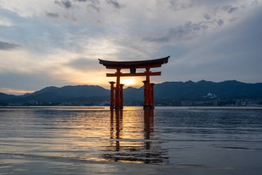 Itsukushima jinja Tapınağı 'nın ünlü yüzen torii kapısı Miyajima Adası yakınlarında sakin suları ve çevresini çevreleyen yeşil tepeleri olan günbatımında. Japonya, Hiroşima yakınlarında.