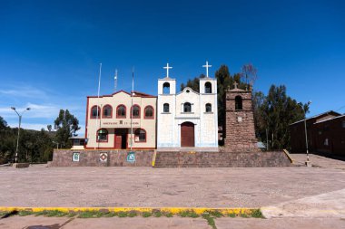 Peru 'nun Llachon kentindeki kilise ve belediye binası, arka planda açık mavi bir gökyüzü ve ağaçlarla birlikte bir taş plazanın üzerinde yan yana duruyor.