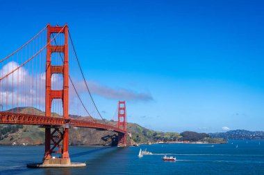 Golden Gate Köprüsü körfez boyunca uzanır, kırmızı kuleleri mavi gökyüzü ve su ile zıtlaşır, arka planda yeşil tepeler vardır.