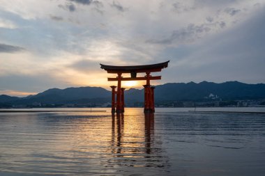 Itsukushima jinja Tapınağı 'nın ünlü yüzen torii kapısı Miyajima Adası yakınlarında sakin suları ve çevresini çevreleyen yeşil tepeleri olan günbatımında. Japonya, Hiroşima yakınlarında.