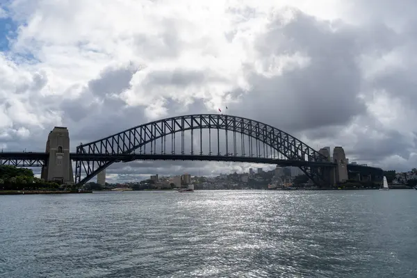Sydney, Avustralya 'daki Sydney Harbour Köprüsü bulutlu bir gökyüzünün altında liman boyunca uzanır. Çelik kemer köprüsü şehri birbirine bağlıyor, etkileyici yapısının altında yüzen teknelerle gökyüzü ve su manzarası sunuyor.