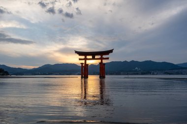 Itsukushima jinja Tapınağı 'nın ünlü yüzen torii kapısı Miyajima Adası yakınlarında sakin suları ve çevresini çevreleyen yeşil tepeleri olan günbatımında. Japonya, Hiroşima yakınlarında.