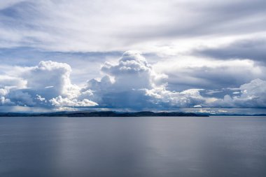 Taquile Adası 'ndan Titicaca Gölü' nün geniş manzarası. Durgun sular, dalgalanan beyaz ve mavi bulutlarla dolu dramatik bir gökyüzünü yansıtıyor.
