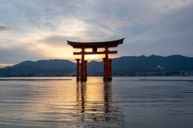 Itsukushima jinja Tapınağı 'nın ünlü yüzen torii kapısı Miyajima Adası yakınlarında sakin suları ve çevresini çevreleyen yeşil tepeleri olan günbatımında. Japonya, Hiroşima yakınlarında.
