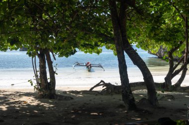 Geleneksel ahşap bir tekne, Lia sahilinin beyaz kumları yakınında, Waleakodi Adası, Togian takımadaları, Sulawesi, Endonezya 'da parlak mavi bir gökyüzünün altında yüzmektedir.