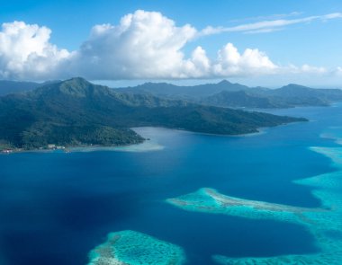 Fransız Polinezyası 'ndaki Tahaa adası, yukarıdan bakıldığında turkuaz bir göl, mercan resifi ve yemyeşil dağları ortaya çıkarır. Canlı okyanus renkleri koyu mavi Pasifik ile tezat oluşturuyor.