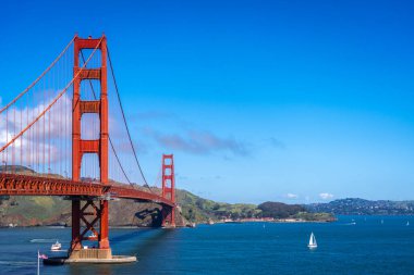 Golden Gate Köprüsü körfez boyunca uzanır, kırmızı kuleleri mavi gökyüzü ve su ile zıtlaşır, arka planda yeşil tepeler vardır.