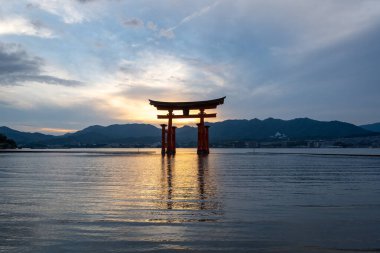 Itsukushima jinja Tapınağı 'nın ünlü yüzen torii kapısı Miyajima Adası yakınlarında sakin suları ve çevresini çevreleyen yeşil tepeleri olan günbatımında. Japonya, Hiroşima yakınlarında.