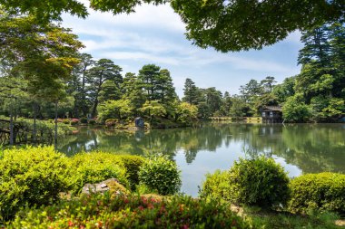 Kanazawa 'daki Kenroku-en Bahçesi tarihi Kanazawa Kalesi' nin yanında huzurlu bir su, yeşillik ve geleneksel yapılar manzarası sunuyor.