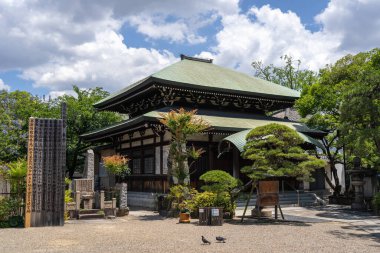 Osaka 'daki Isshin-ji Tapınağı' nın ana salonu parçalı bulutlu bir gökyüzünün altında yontulmuş ağaçlar ve taş fenerler tarafından çerçevelenmiştir.