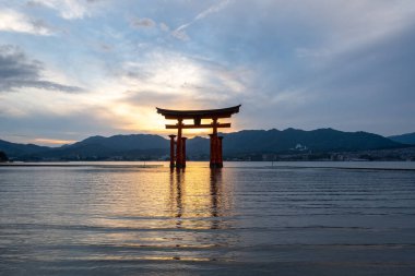 Itsukushima jinja Tapınağı 'nın ünlü yüzen torii kapısı Miyajima Adası yakınlarında sakin suları ve çevresini çevreleyen yeşil tepeleri olan günbatımında. Japonya, Hiroşima yakınlarında.
