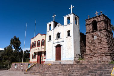 Peru 'nun Llachon kentindeki kilise ve belediye binası, arka planda açık mavi bir gökyüzü ve ağaçlarla birlikte bir taş plazanın üzerinde yan yana duruyor.
