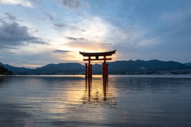 Itsukushima jinja Tapınağı 'nın ünlü yüzen torii kapısı Miyajima Adası yakınlarında sakin suları ve çevresini çevreleyen yeşil tepeleri olan günbatımında. Japonya, Hiroşima yakınlarında.