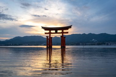 Itsukushima jinja Tapınağı 'nın ünlü yüzen torii kapısı Miyajima Adası yakınlarında sakin suları ve çevresini çevreleyen yeşil tepeleri olan günbatımında. Japonya, Hiroşima yakınlarında.