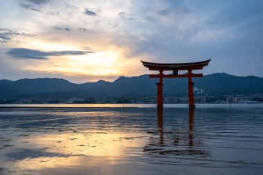 Itsukushima jinja Tapınağı 'nın ünlü yüzen torii kapısı Miyajima Adası yakınlarında sakin suları ve çevresini çevreleyen yeşil tepeleri olan günbatımında. Japonya, Hiroşima yakınlarında.