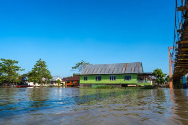Renkli bir asma köprü, Endonezya, Sulawesi 'de canlı mavi bir gökyüzü altında nehir kenarındaki evleri birbirine bağlayan Tempe Gölü' nün sakin sularını kaplar.