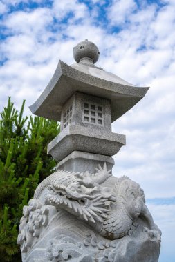 Japonya 'nın Enoshima adasındaki parlak bir gökyüzünün altında, efsanevi sanatla geleneksel tasarımı harmanlayan bir ejderha heykeli bulunan süslemeli taş fener.