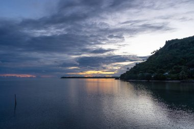 Altın bir günbatımı, Fransız Polinezyası 'ndaki Maupiti gölünün sakin sularını dramatik bulutlar ve kıyı şeridi boyunca uzanan siluetli palmiye ağaçlarıyla yansıtır.